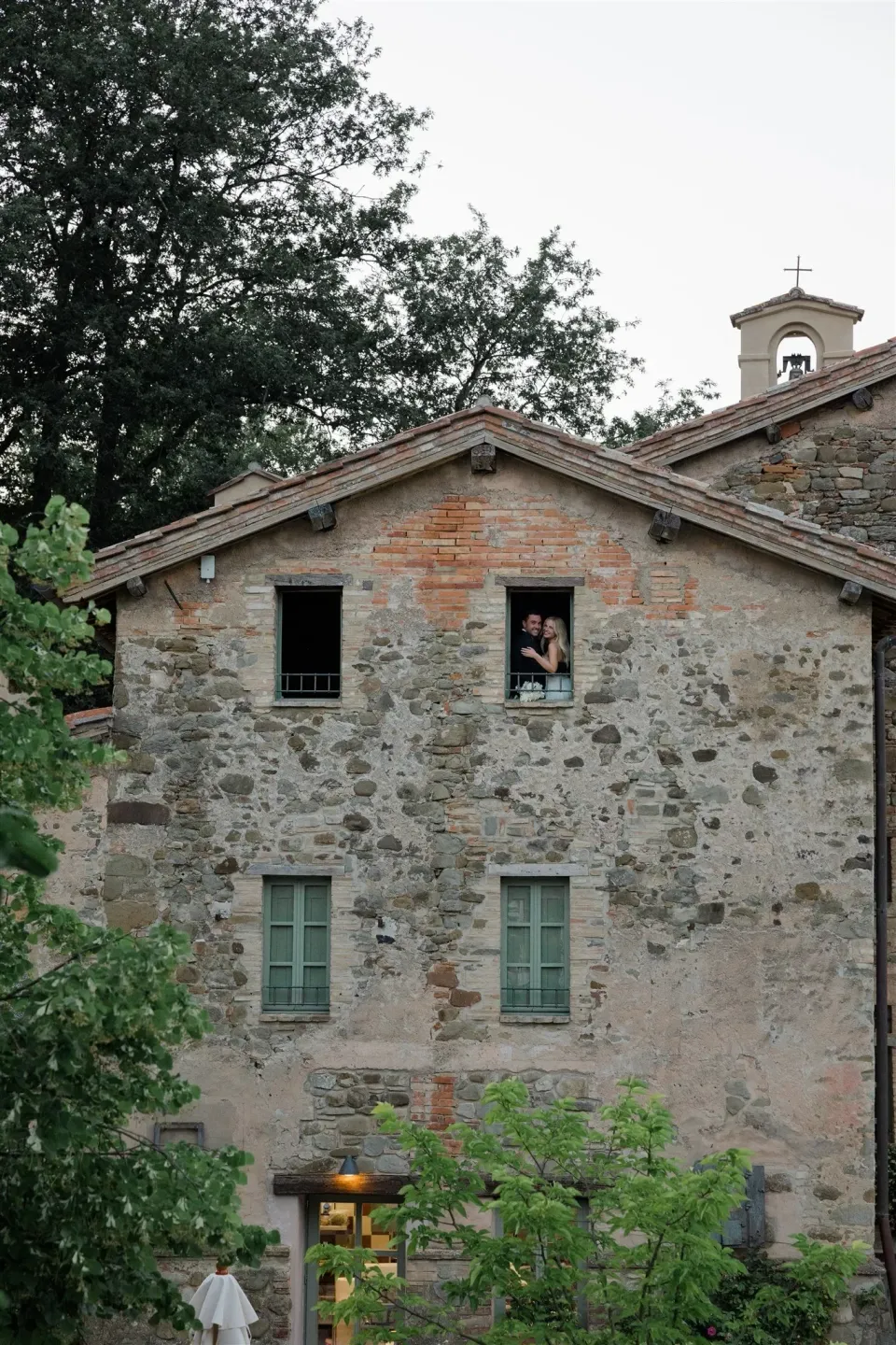 Shelby and Jack looking out from an upstairs window in a stone country building, in Umbria, Italy, for an Elopement Collective elopement. Photo by James from House of Love.