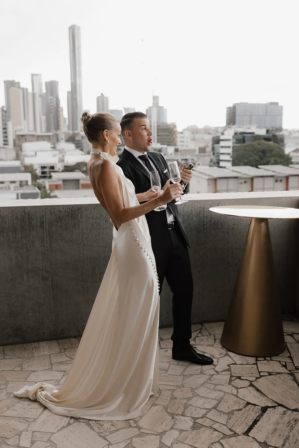 Clare and Matt holding champagne on a terrace with the city skyline behind them, at The Calile Hotel in Fortitude Valley, Brisbane, for an Elopement Collective elopement. Photo by Harley from Bulb Creative.