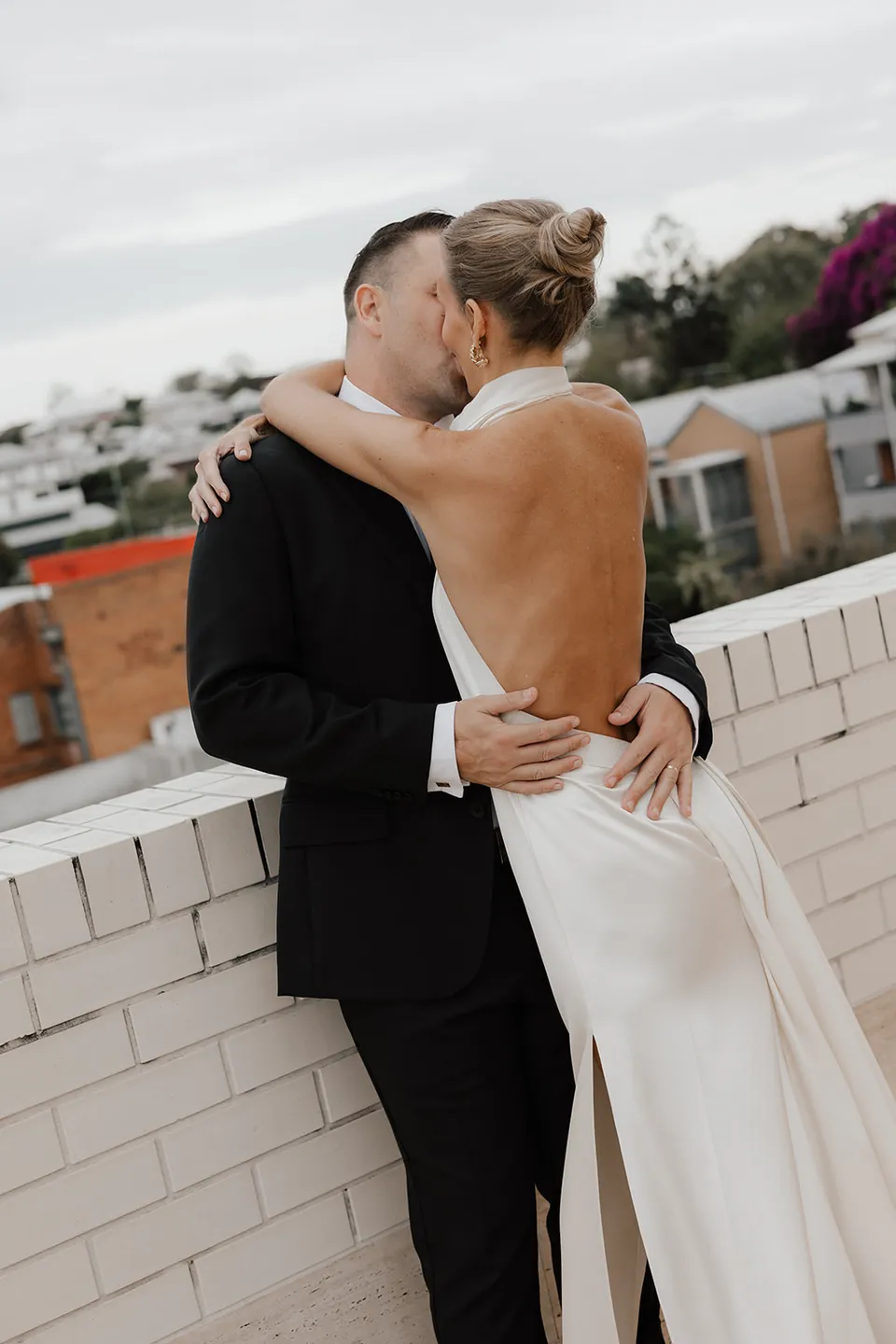 Clare and Matt embracing beside a white brick terrace wall, at The Calile Hotel in Fortitude Valley, Brisbane, for an Elopement Collective elopement. Photo by Harley from Bulb Creative.