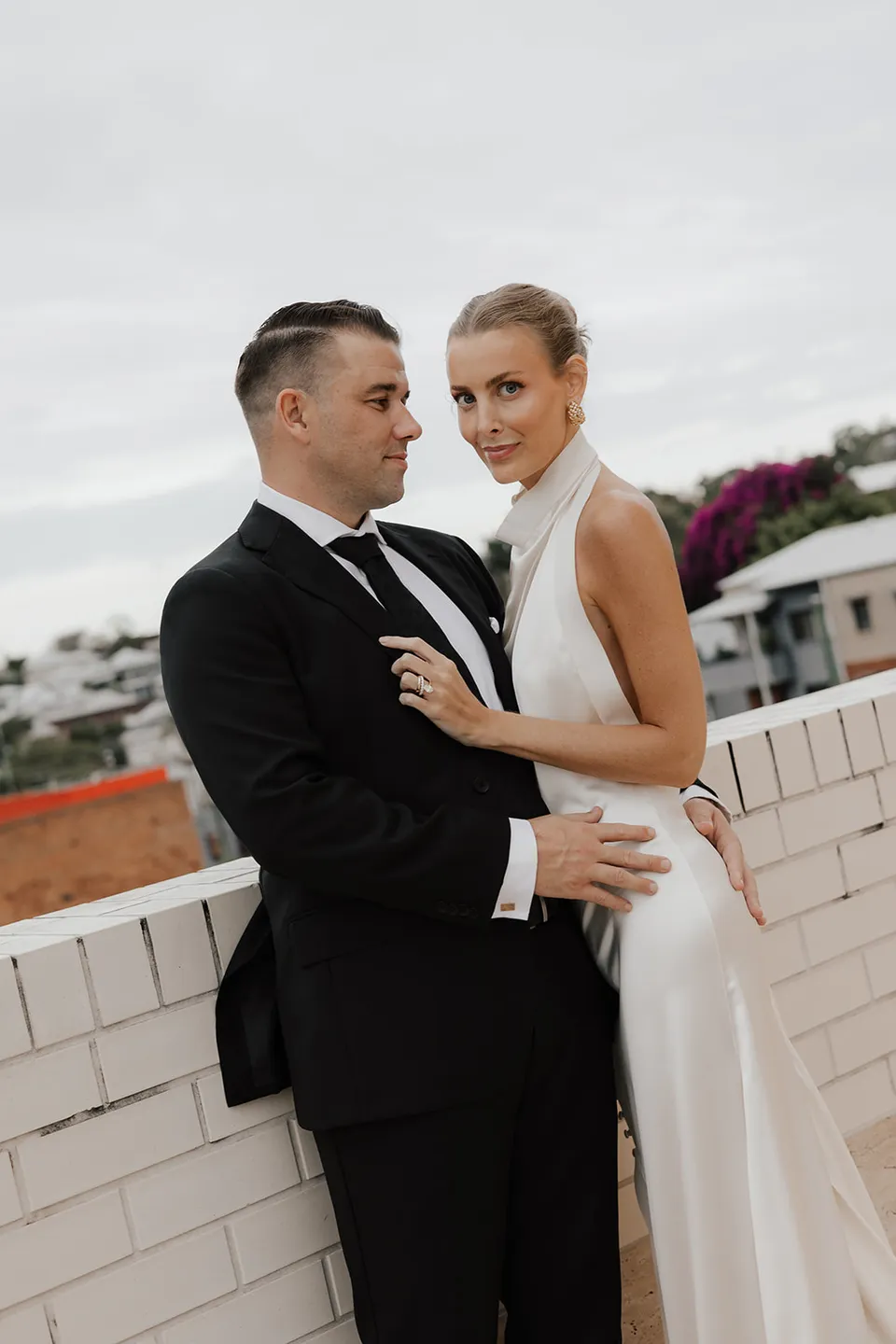 Clare and Matt standing together by a white brick wall with the city behind them, at The Calile Hotel in Fortitude Valley, Brisbane, for an Elopement Collective elopement. Photo by Harley from Bulb Creative.