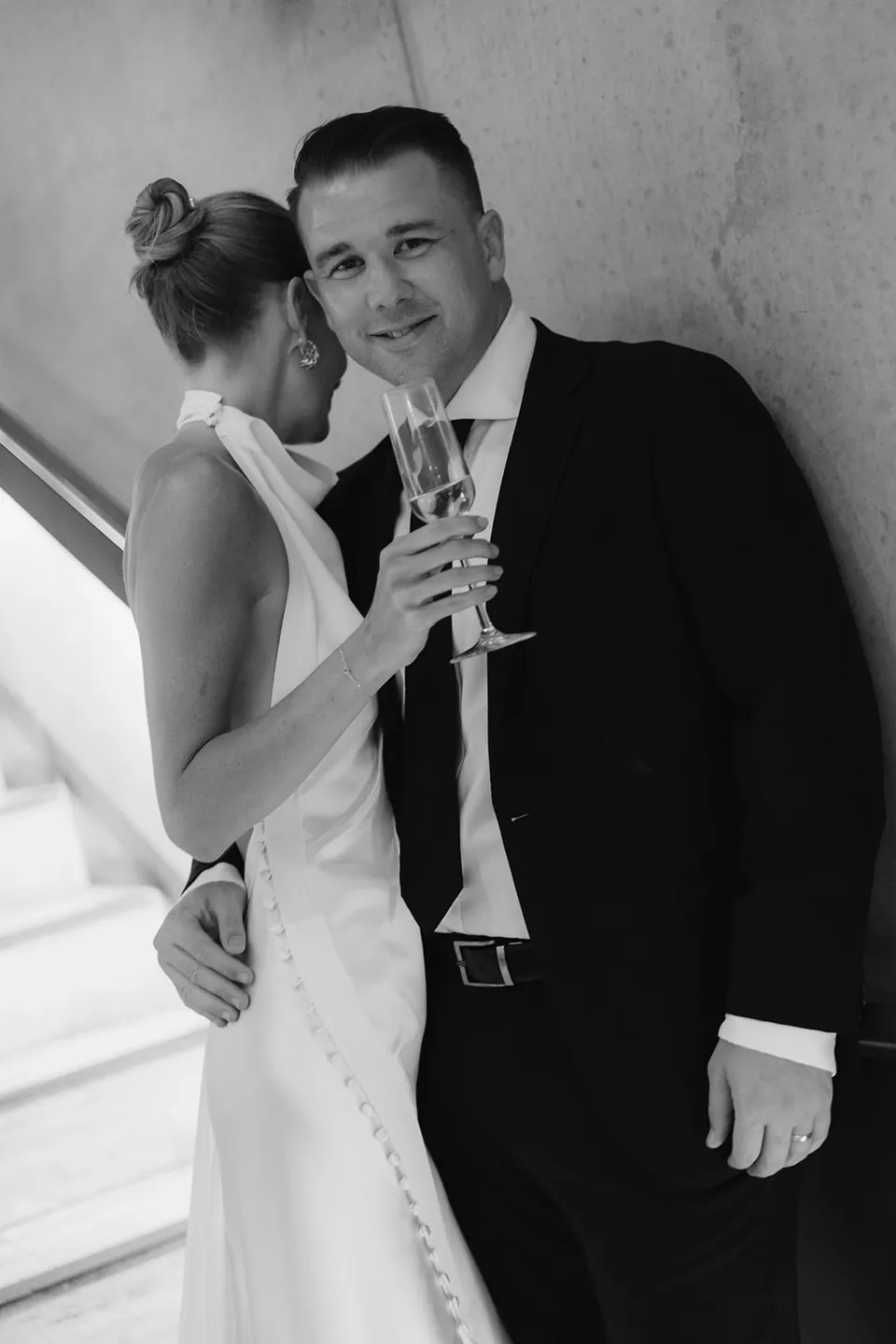 Black-and-white portrait of Clare leaning into Matt while holding champagne, at The Calile Hotel in Fortitude Valley, Brisbane, for an Elopement Collective elopement. Photo by Harley from Bulb Creative.