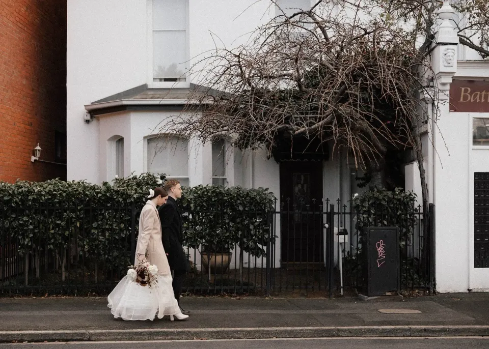 A couple walk past a white cottage, bare branches and an iron fence.