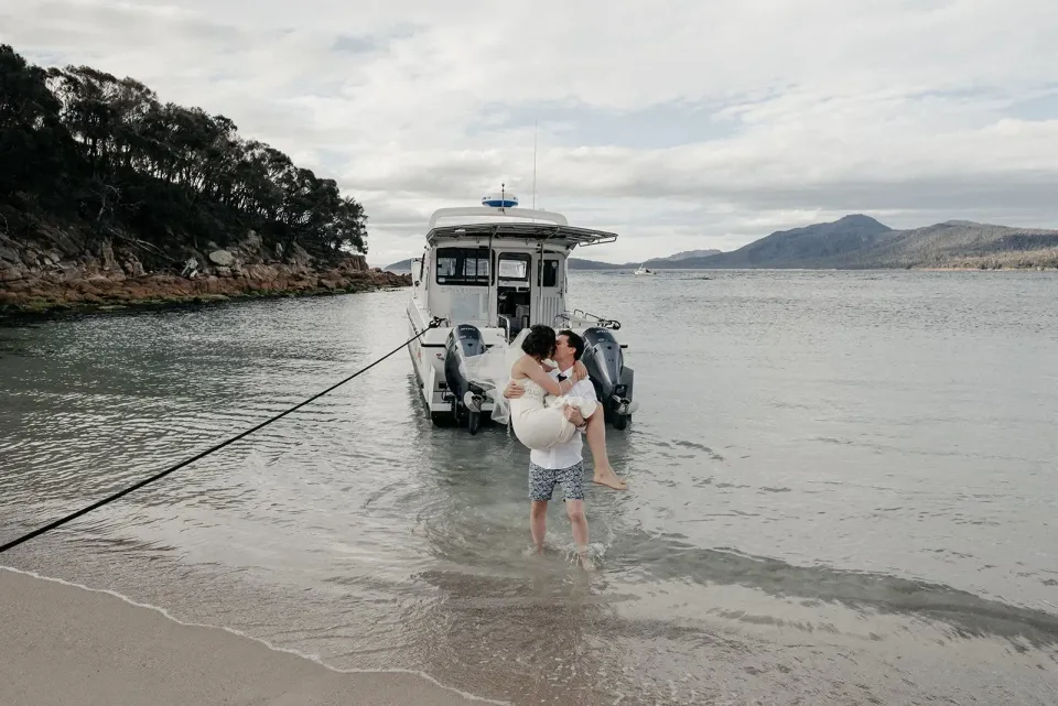 A groom carries his bride through shallow water beside a boat at the beach.