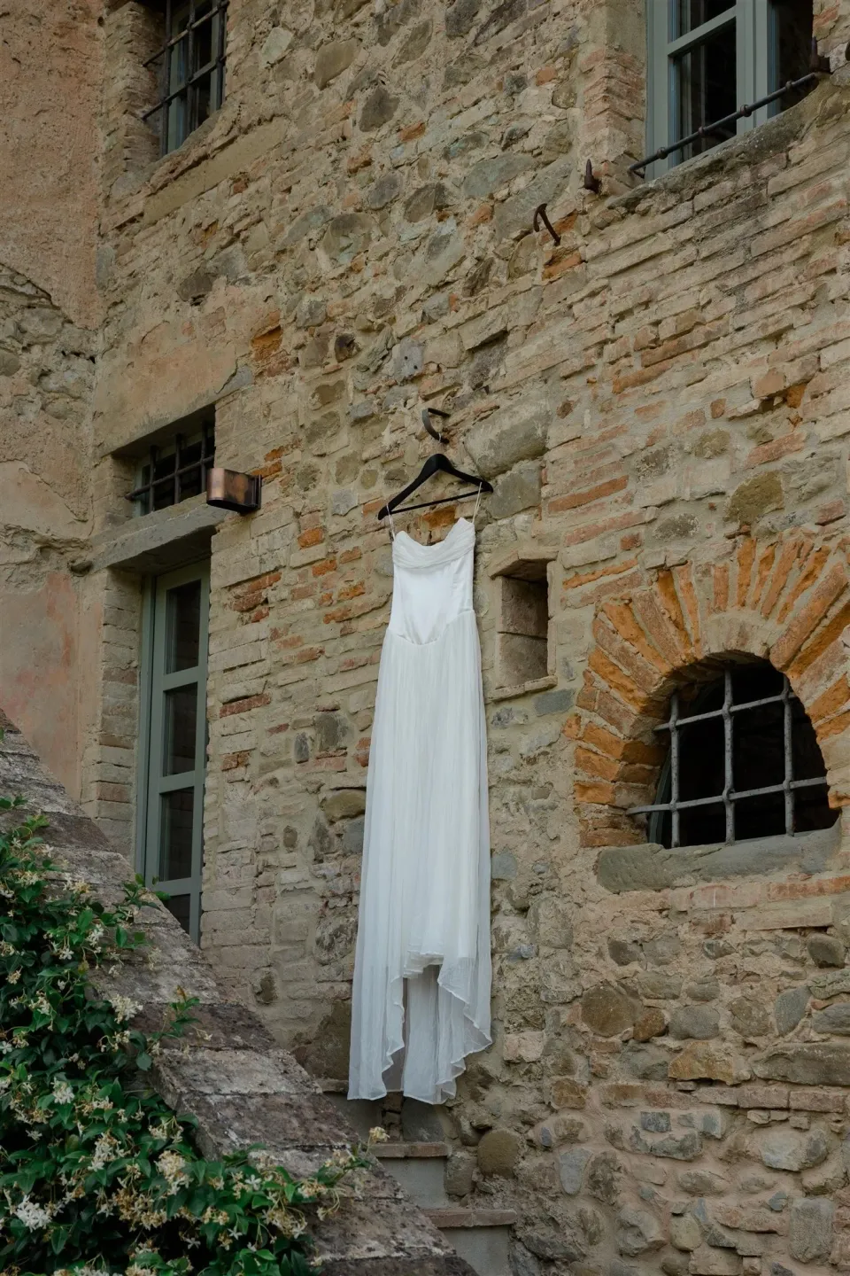 Shelby’s white Kate Willa wedding dress hanging against a rustic stone wall, in Umbria, Italy, for an Elopement Collective elopement. Photo by James from House of Love.