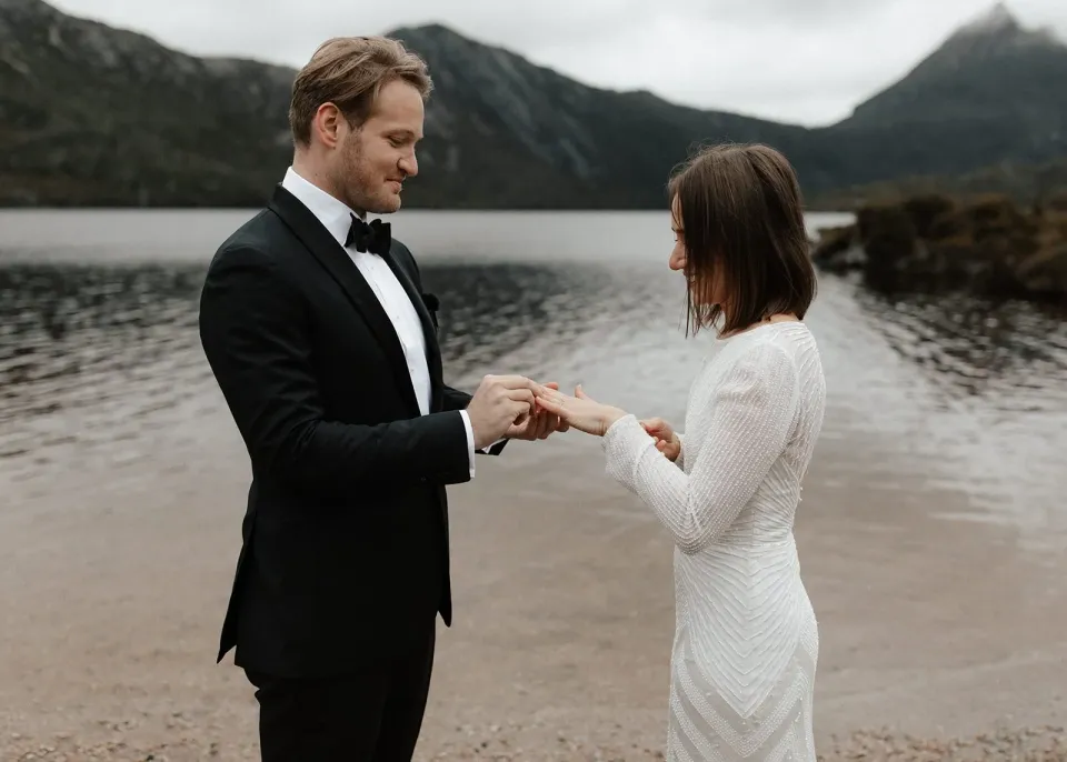 A couple exchange rings beside a mountain lake.