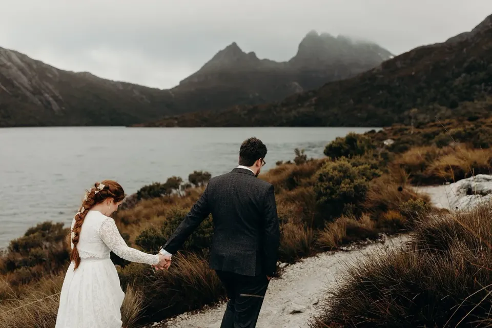 A couple walk hand in hand beside a lake beneath rugged mountain peaks.