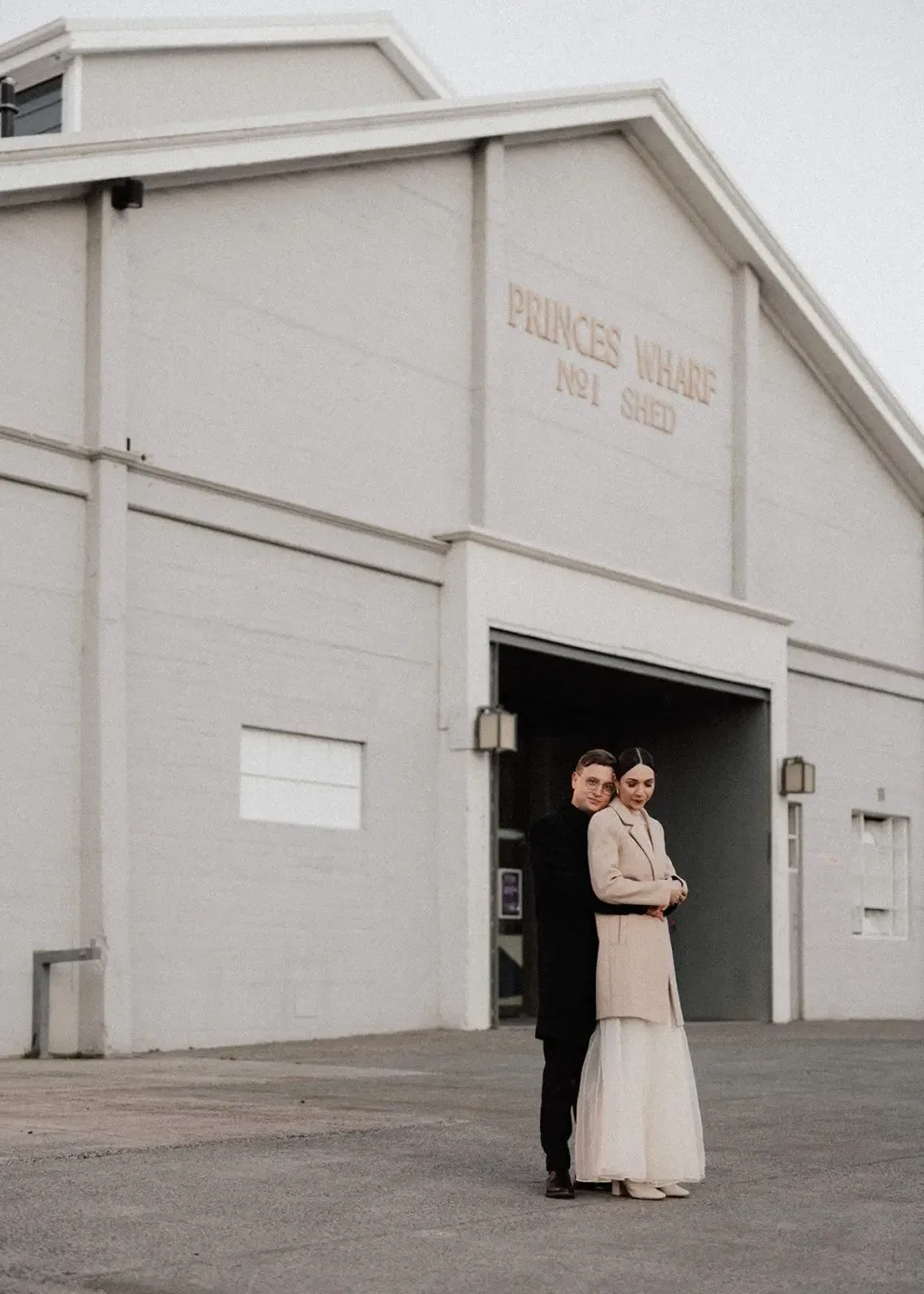 A couple embrace outside the open doorway of a pale waterfront shed.