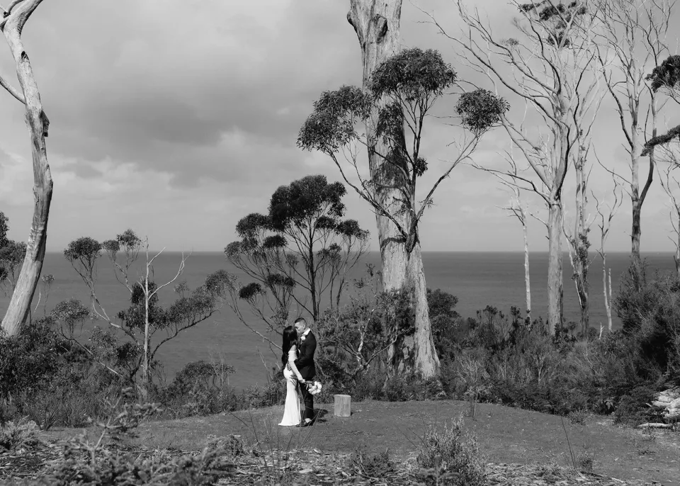 A black-and-white photograph of a couple beneath tall gum trees overlooking the water.