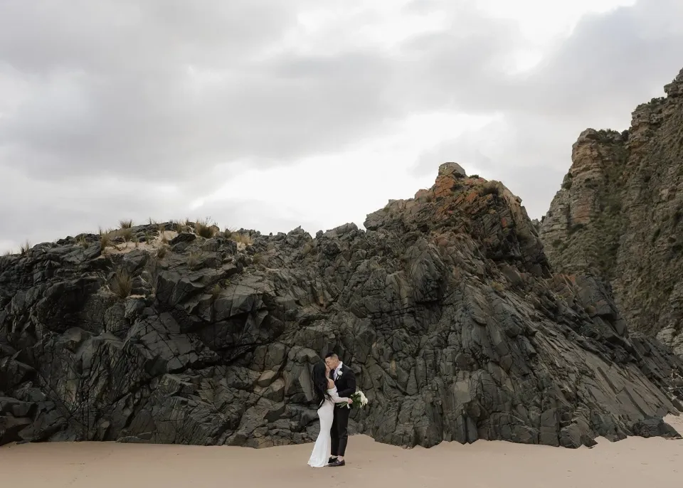 A couple stand together on a beach in front of a rugged wall of coastal rock.