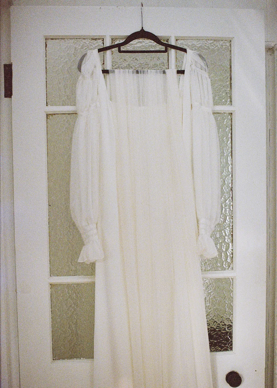 On 35mm film: Elyse's white dress hanging against the glass-panelled bedroom door. At Fig Tree Villa in Ewingsdale, Byron Bay, for an Elopement Collective elopement. Photograph by Bulb Creative.