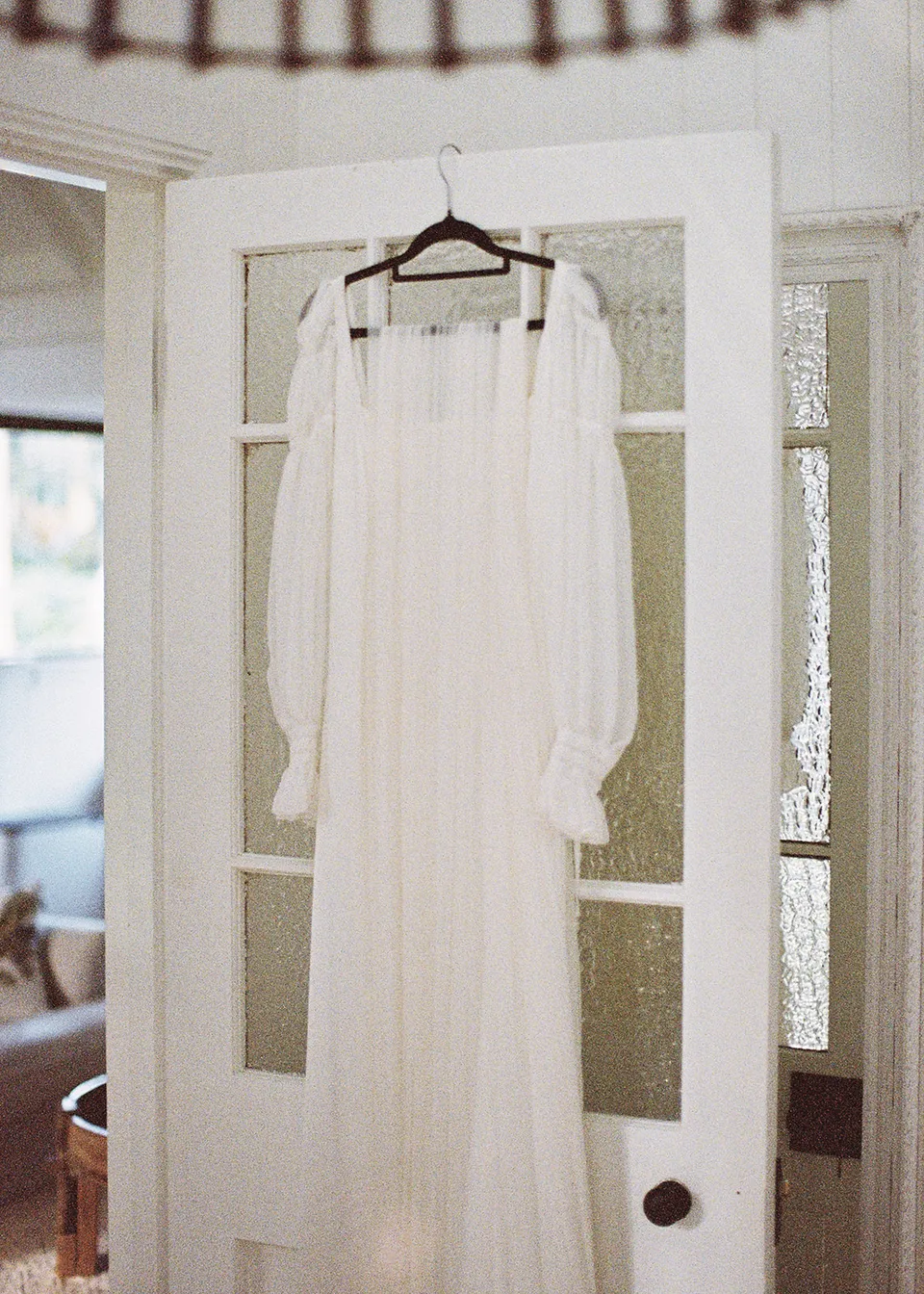 On 35mm film: Elyse's wedding dress hanging in the open doorway, its sheer sleeves catching the light. At Fig Tree Villa in Ewingsdale, Byron Bay, for an Elopement Collective elopement. Photograph by Bulb Creative.