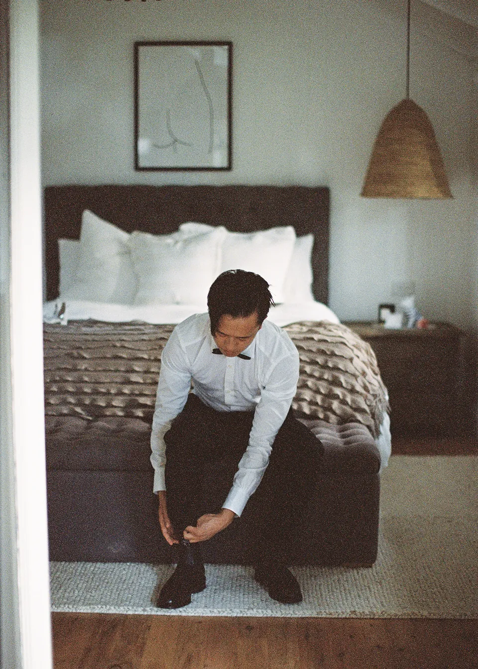 On 35mm film: Jonathan leaning forward to fasten his boots beside the bed. At Fig Tree Villa in Ewingsdale, Byron Bay, for an Elopement Collective elopement. Photograph by Bulb Creative.