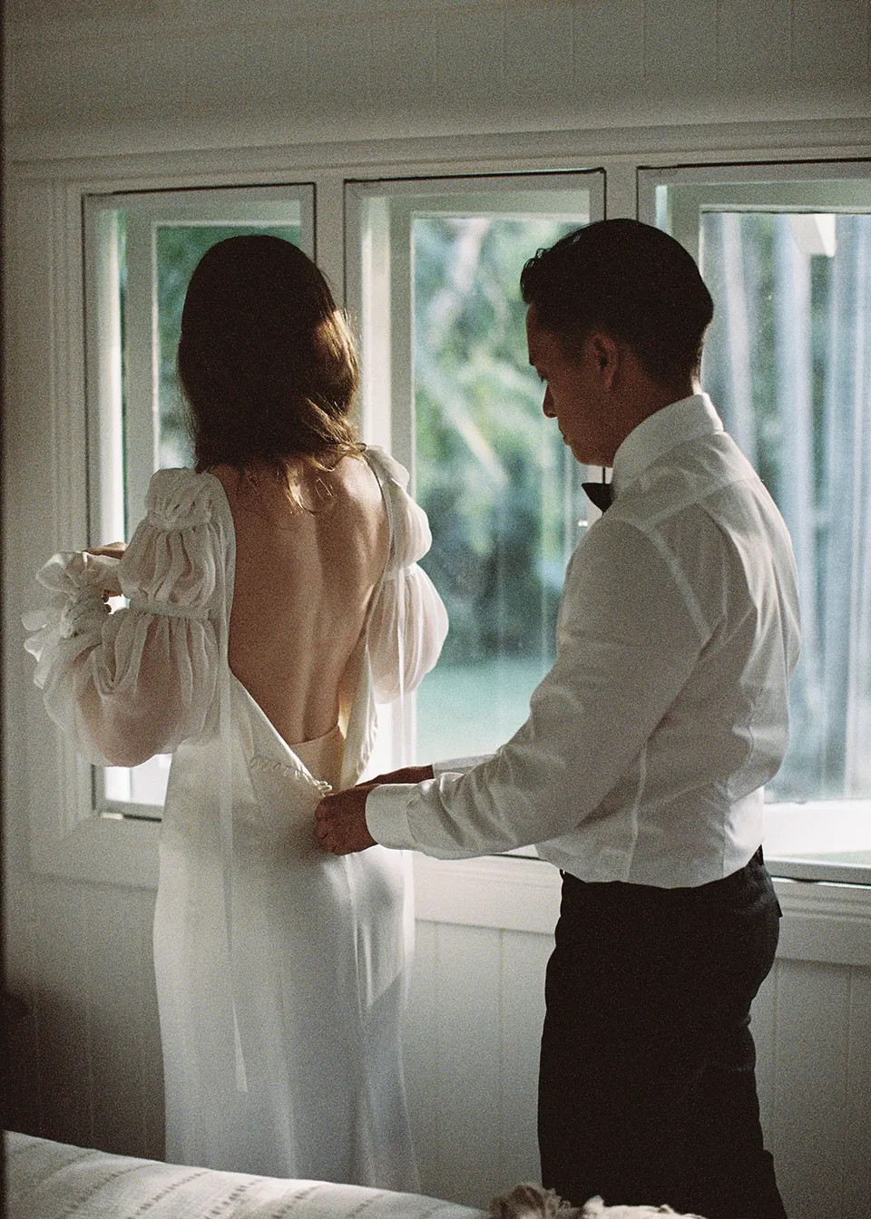 On 35mm film: Jonathan helping fasten Elyse's dress beside the window. At Fig Tree Villa in Ewingsdale, Byron Bay, for an Elopement Collective elopement. Photograph by Bulb Creative.