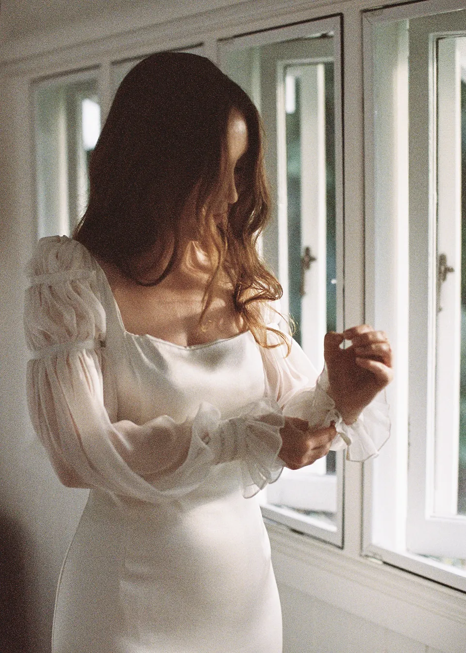 On 35mm film: Elyse looking down while fastening her cuff beside the window. At Fig Tree Villa in Ewingsdale, Byron Bay, for an Elopement Collective elopement. Photograph by Bulb Creative.