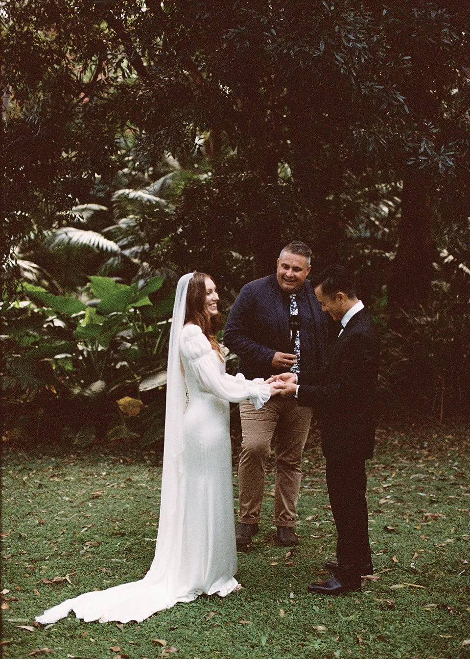 On 35mm film: The couple listening to celebrant Josh Withers beneath the leafy canopy. At Fig Tree Villa in Ewingsdale, Byron Bay, for an Elopement Collective elopement. Photograph by Bulb Creative.