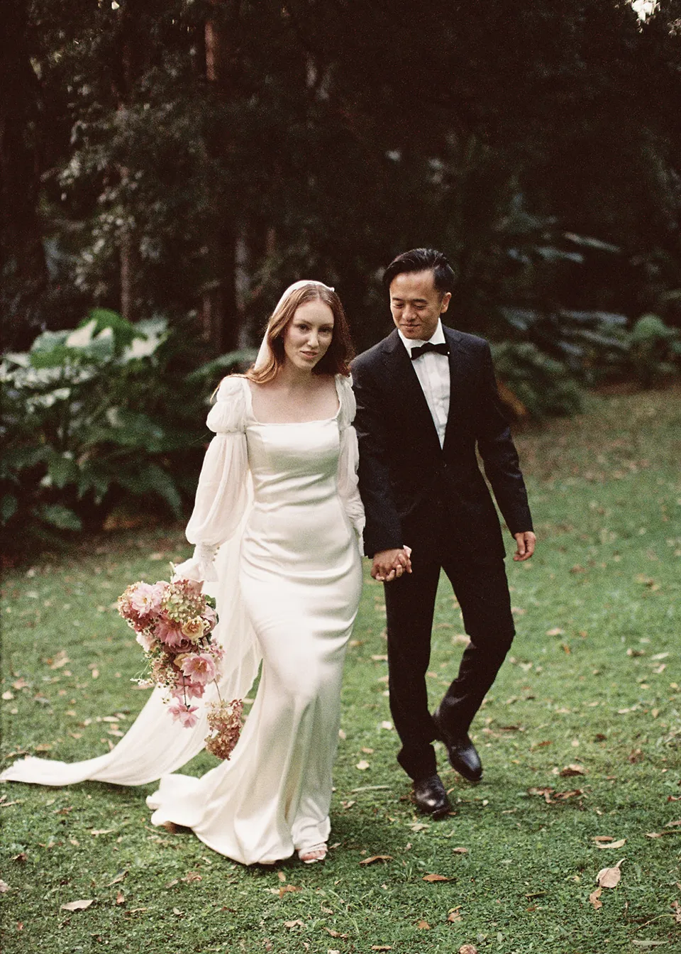 On 35mm film: Elyse and Jonathan walking towards the camera beneath the leafy canopy. At Fig Tree Villa in Ewingsdale, Byron Bay, for an Elopement Collective elopement. Photograph by Bulb Creative.