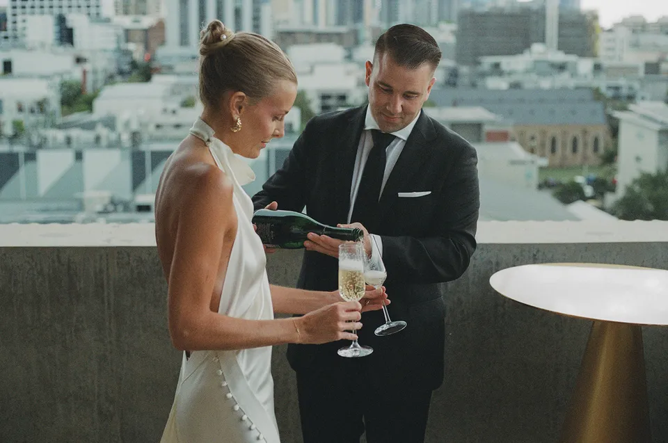 Matt reading from a green vow booklet beside Clare on the hotel terrace, at The Calile Hotel in Fortitude Valley, Brisbane, for an Elopement Collective elopement. Photo by Harley from Bulb Creative.