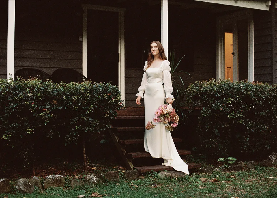 On 35mm film: Wide portrait of Elyse beside the cottage verandah and garden hedge. At Fig Tree Villa in Ewingsdale, Byron Bay, for an Elopement Collective elopement. Photograph by Bulb Creative.