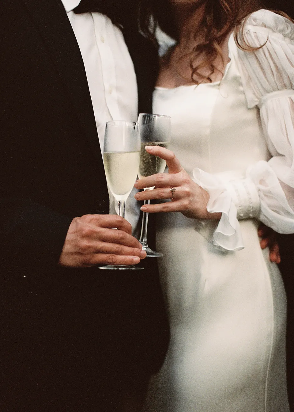 On 35mm film: The couple's hands around their glasses beside Elyse's white dress. At Fig Tree Villa in Ewingsdale, Byron Bay, for an Elopement Collective elopement. Photograph by Bulb Creative.