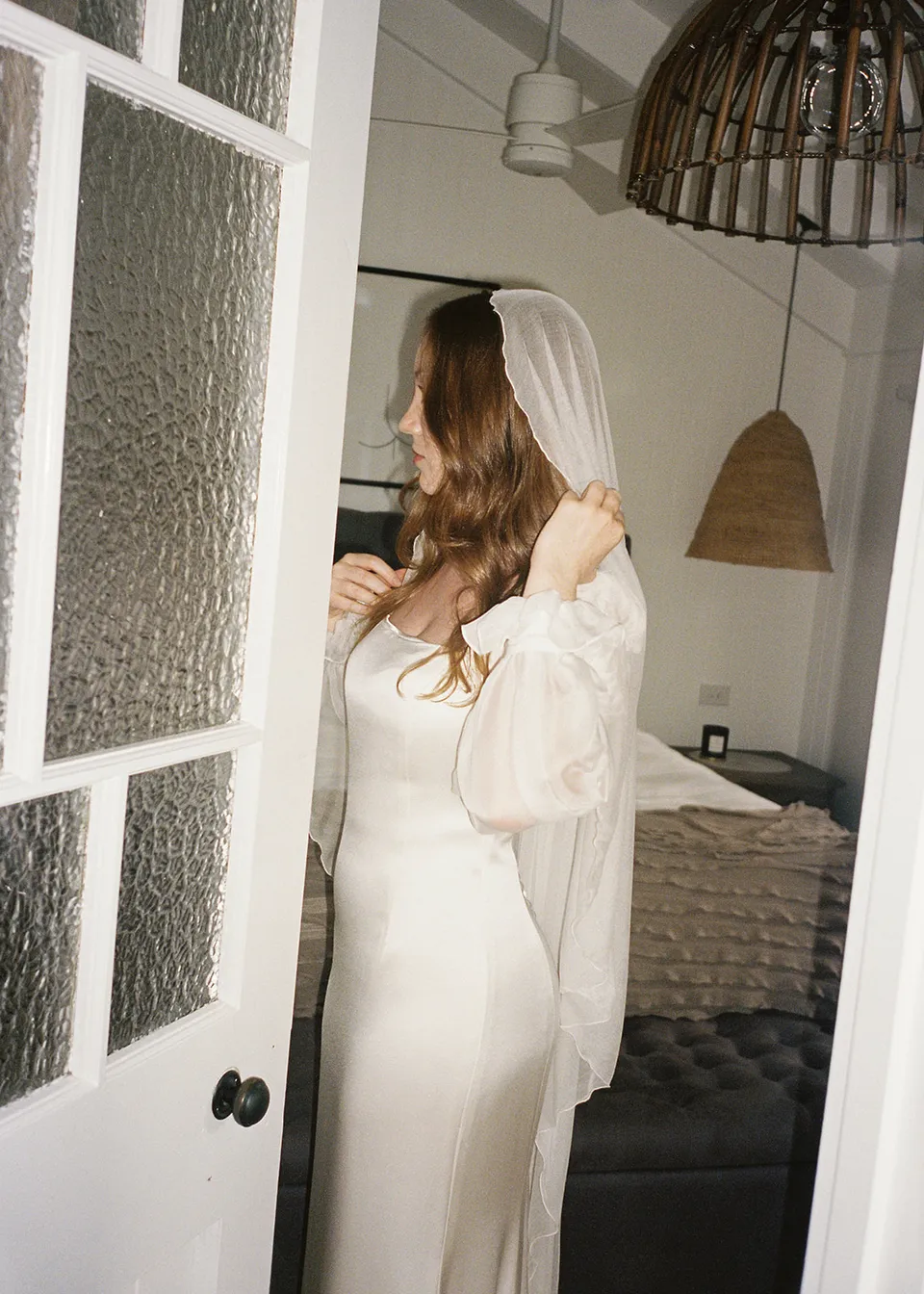 On 35mm film: Elyse standing in her veil at the glass-panelled doorway. At Fig Tree Villa in Ewingsdale, Byron Bay, for an Elopement Collective elopement. Photograph by Bulb Creative.