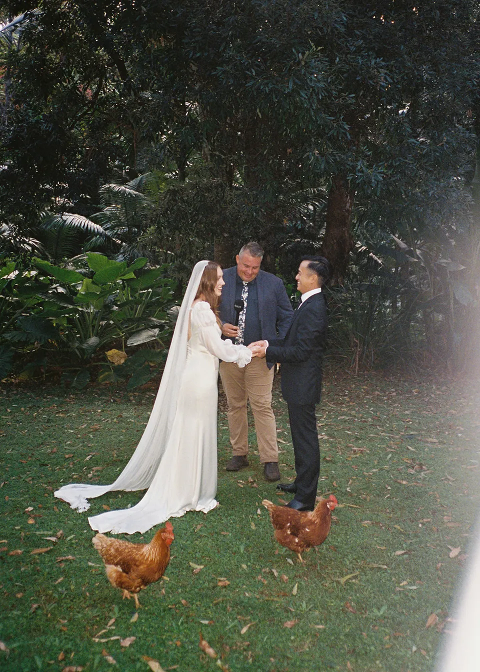 On 35mm film: Brown hens wandering past the couple and celebrant Josh Withers during the garden ceremony. At Fig Tree Villa in Ewingsdale, Byron Bay, for an Elopement Collective elopement. Photograph by Bulb Creative.