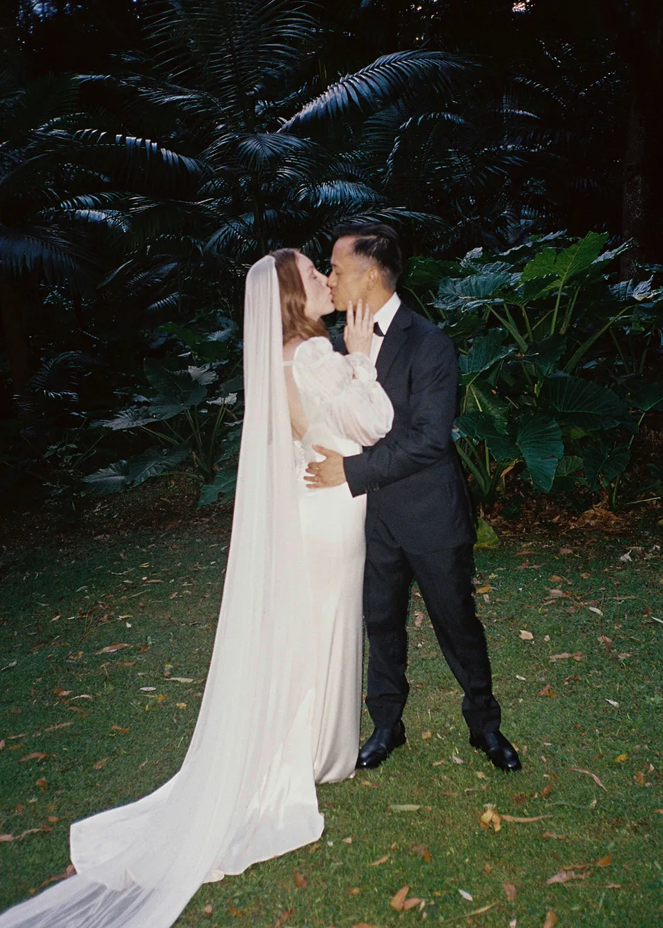 On 35mm film: The couple kissing on the lawn with Elyse's veil trailing behind her. At Fig Tree Villa in Ewingsdale, Byron Bay, for an Elopement Collective elopement. Photograph by Bulb Creative.