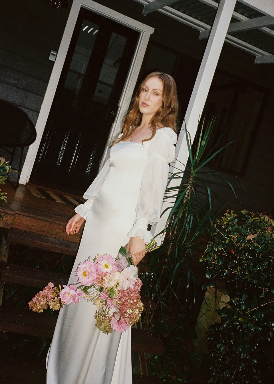 On 35mm film: Elyse holding her bouquet in front of the verandah, with flowers by The Perfect Party Co. At Fig Tree Villa in Ewingsdale, Byron Bay, for an Elopement Collective elopement. Photograph by Bulb Creative.