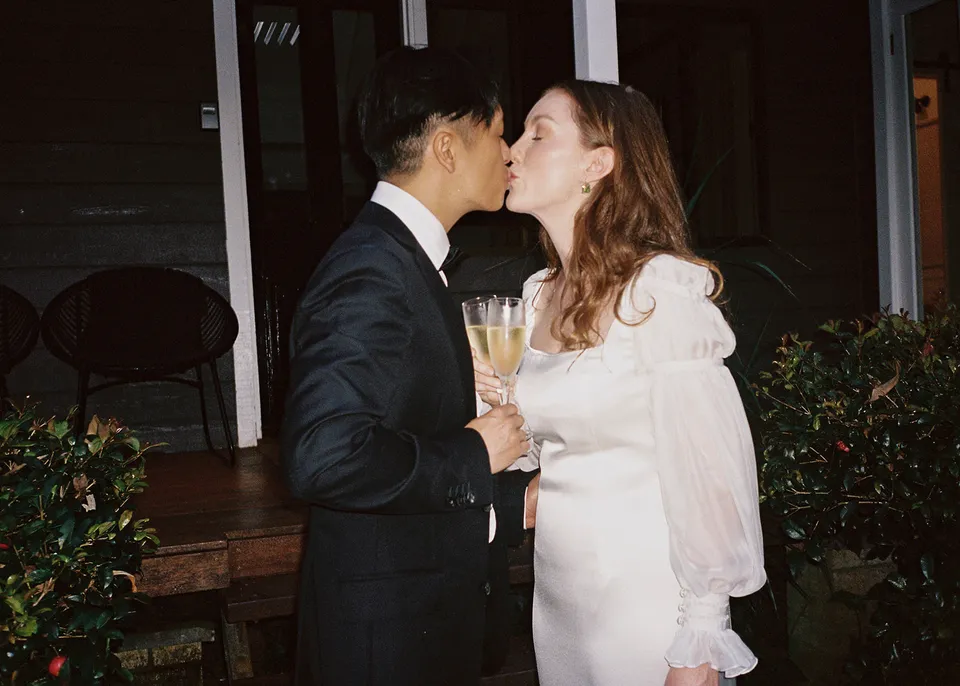 On 35mm film: Elyse and Jonathan kissing outside the cottage with sparkling wine in hand. At Fig Tree Villa in Ewingsdale, Byron Bay, for an Elopement Collective elopement. Photograph by Bulb Creative.