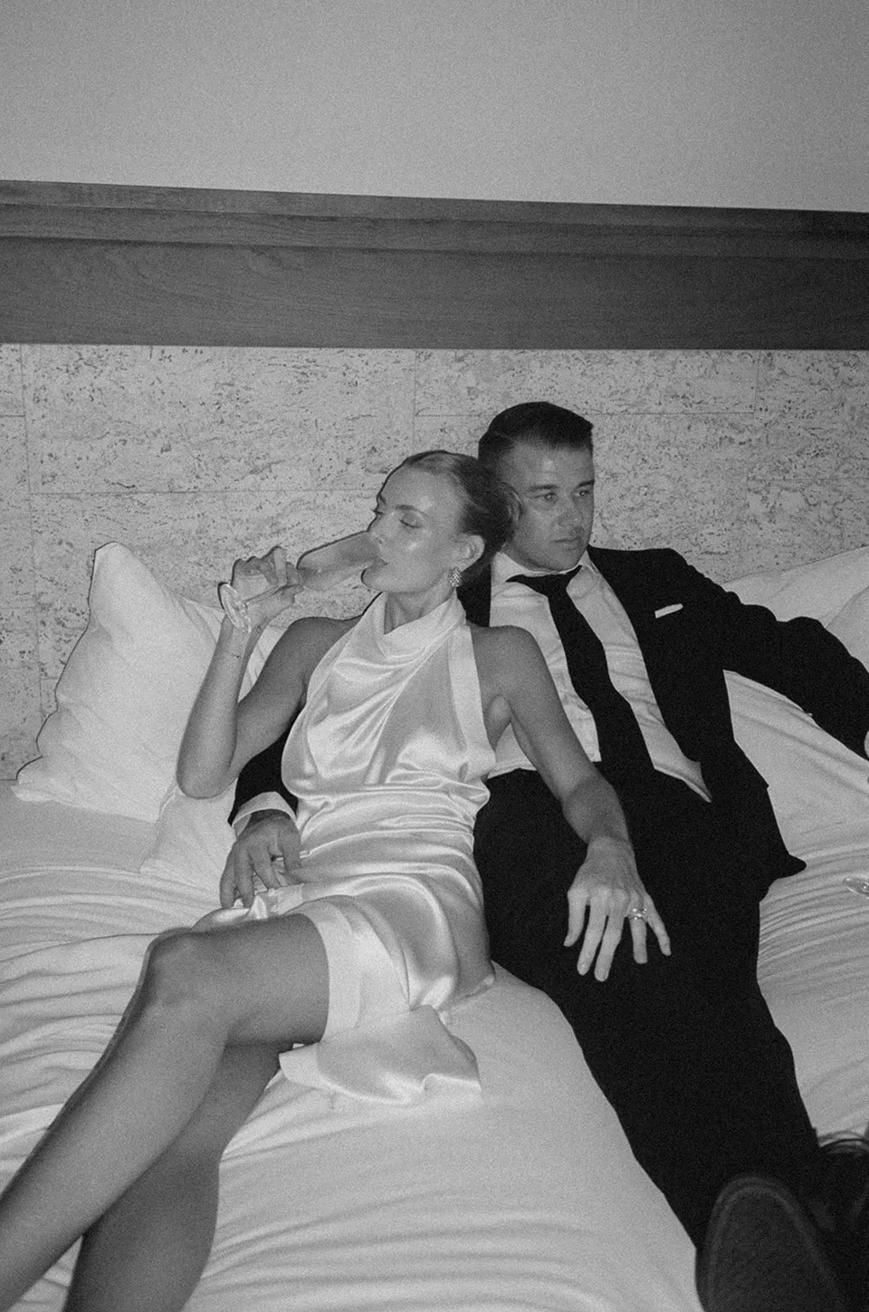 Black-and-white film photograph of Clare sipping champagne beside Matt on a sofa, at The Calile Hotel in Fortitude Valley, Brisbane, for an Elopement Collective elopement. Photo by Harley from Bulb Creative.