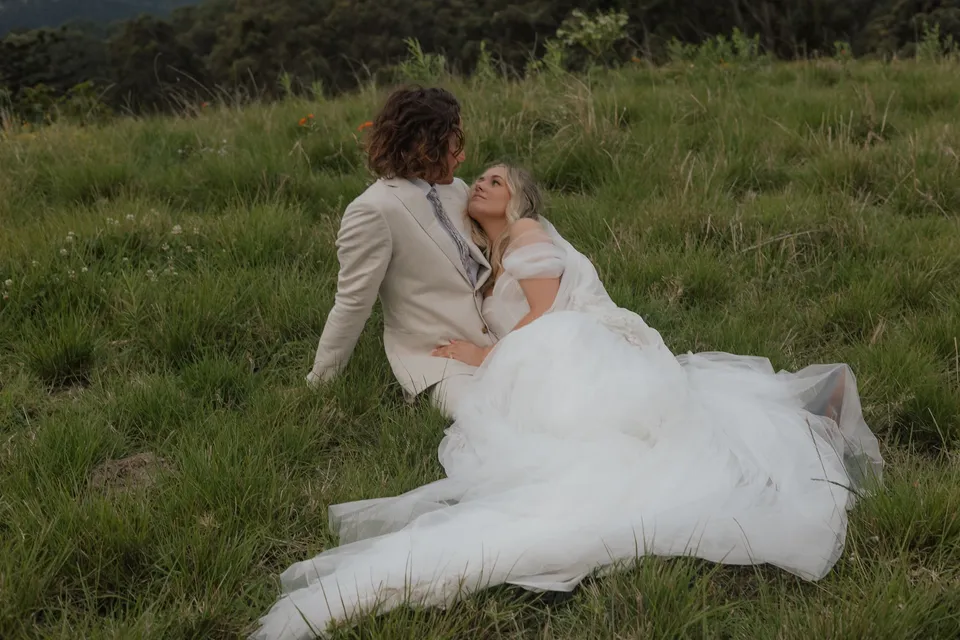 Bridget and James sitting close together in long green grass, her wedding dress spread across the hillside at Cudgera Creek in northern New South Wales. An Elopement Collective elopement, photographed by Bulb Creative.