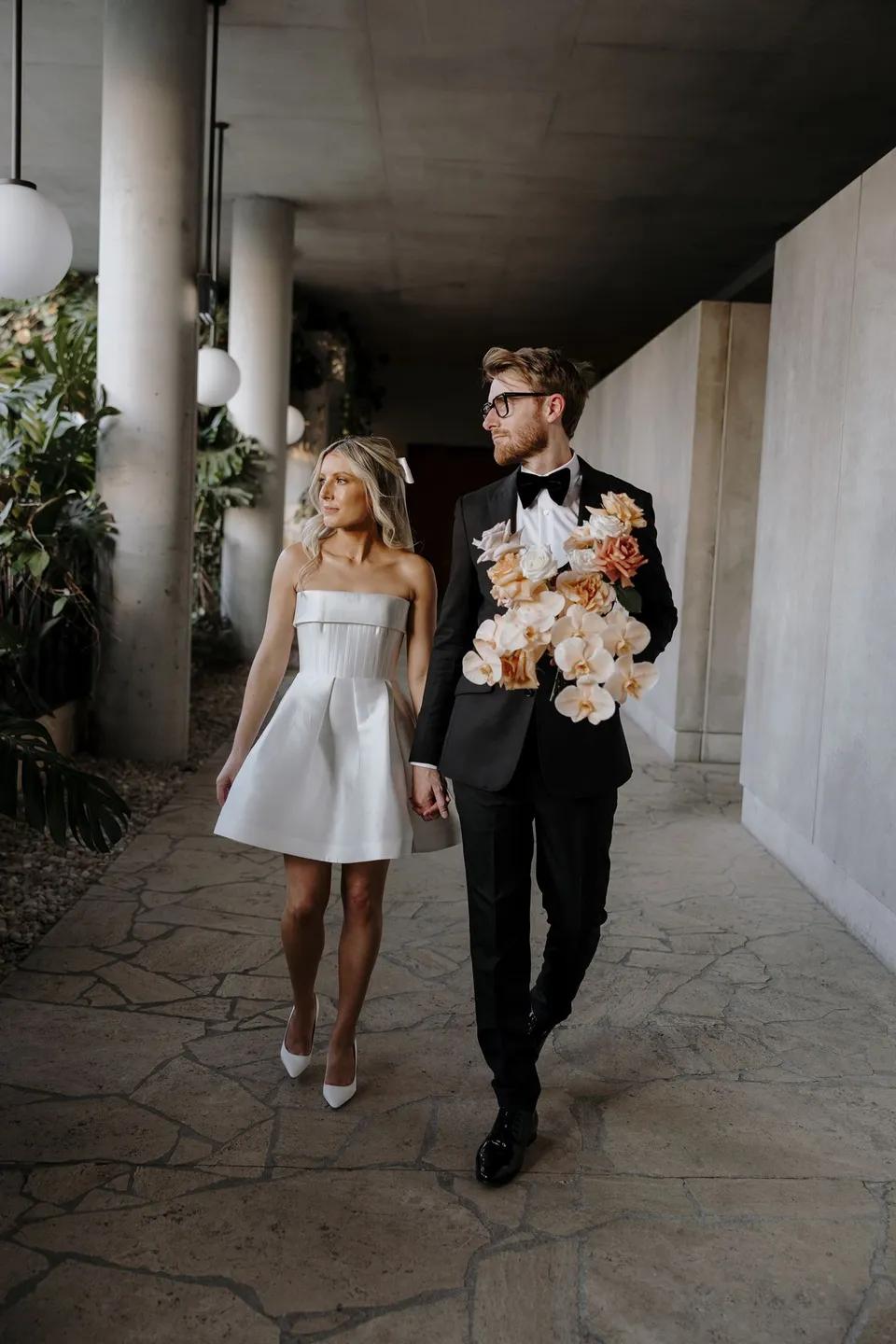 Danielle and Adam walking hand in hand beneath concrete columns, with Adam carrying the peach and white bouquet by Mrs Gibbons Florals, at The Calile Hotel in Fortitude Valley, Brisbane, for an Elopement Collective elopement. Photo by George Bowden.