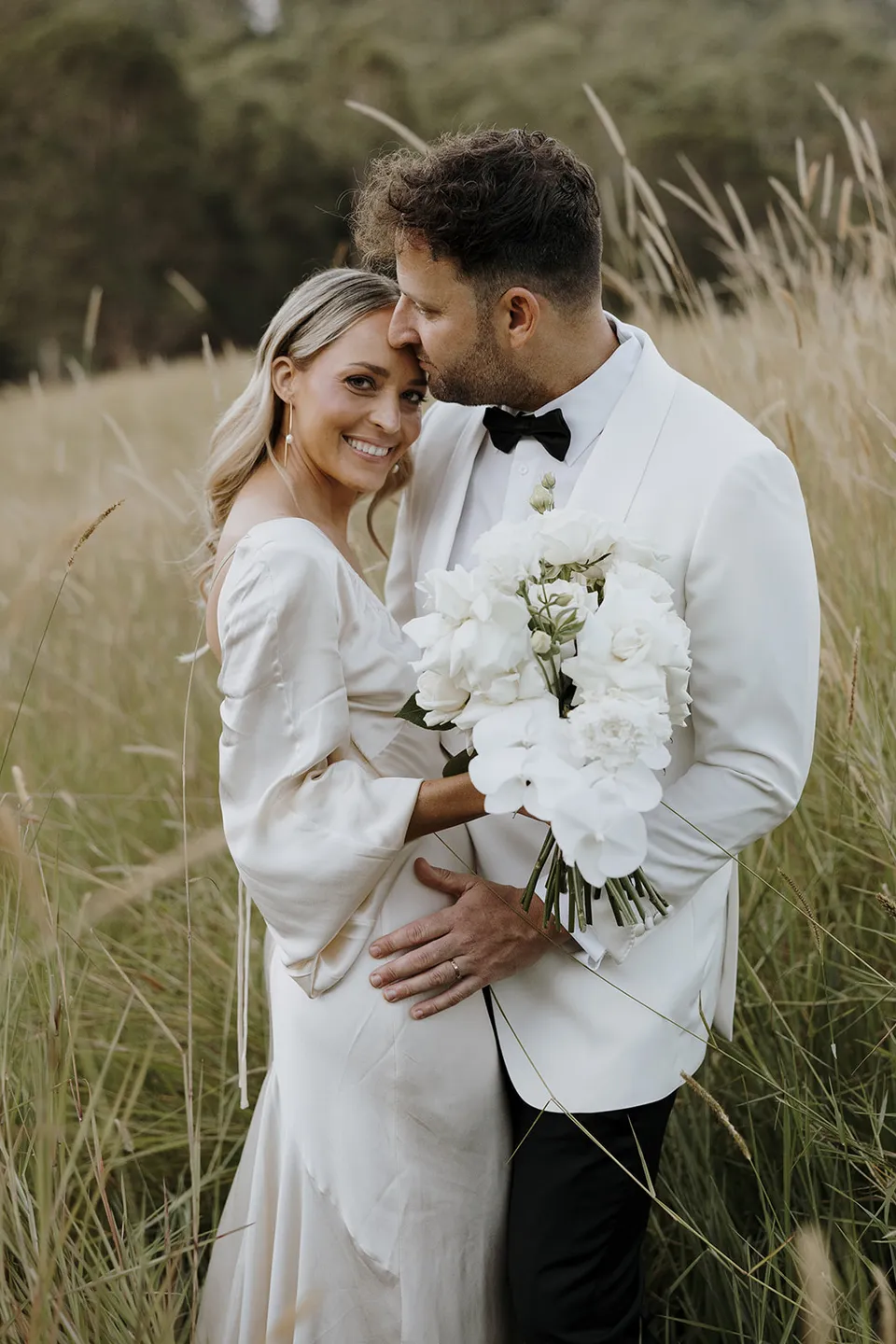 Greg kissing Gabrielle’s forehead as she smiles and holds her white bouquet by Mrs Gibbons in the paddock, at Roundhill Retreat in Obi Obi, Sunshine Coast Hinterland, Queensland, for an Elopement Collective elopement. Photo by George Bowden.