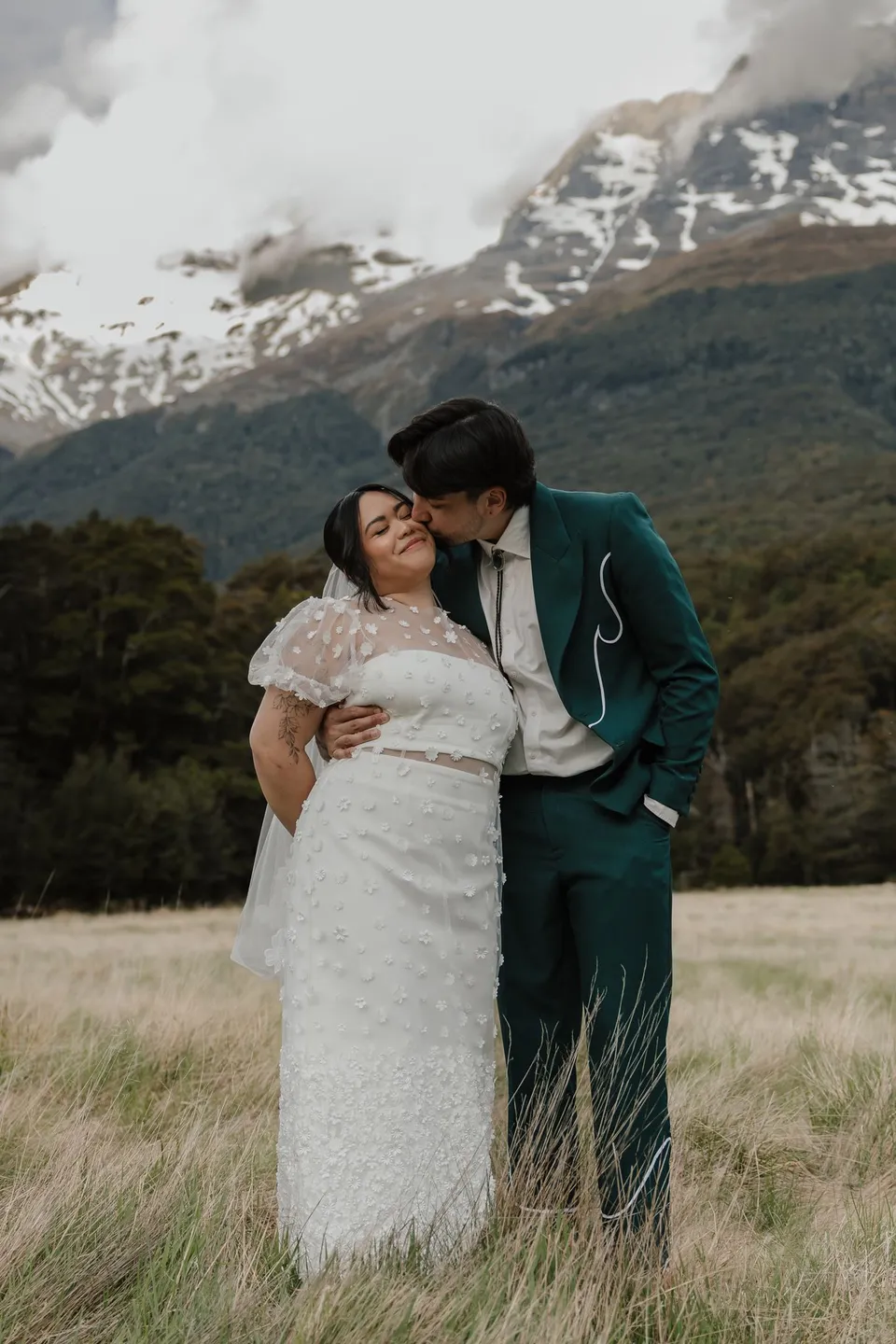 Adrian kissing Jaeda’s cheek as they stand in golden grass beneath snow-streaked mountains, in the field on the road to Paradise near Glenorchy, in the Queenstown region of New Zealand, for an Elopement Collective Queenstown elopement. Photograph by Bulb Creative.