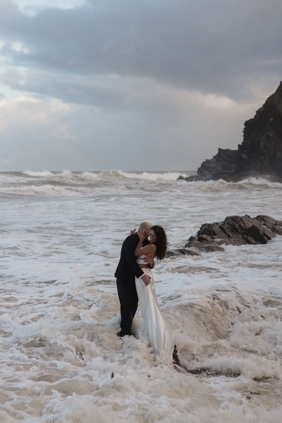 Rachel and Dane embracing in foamy surf below a rocky headland at Whites Beach, Broken Head, near Byron Bay. An Elopement Collective elopement, photographed by Jason Lucas.