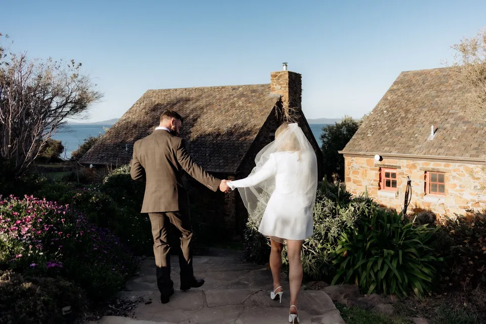 A couple holding hands on a garden path between stone cottages, with slate roofs and blue sea beyond, at Piermont Retreat on Tasmania’s east coast, for an Elopement Collective elopement.