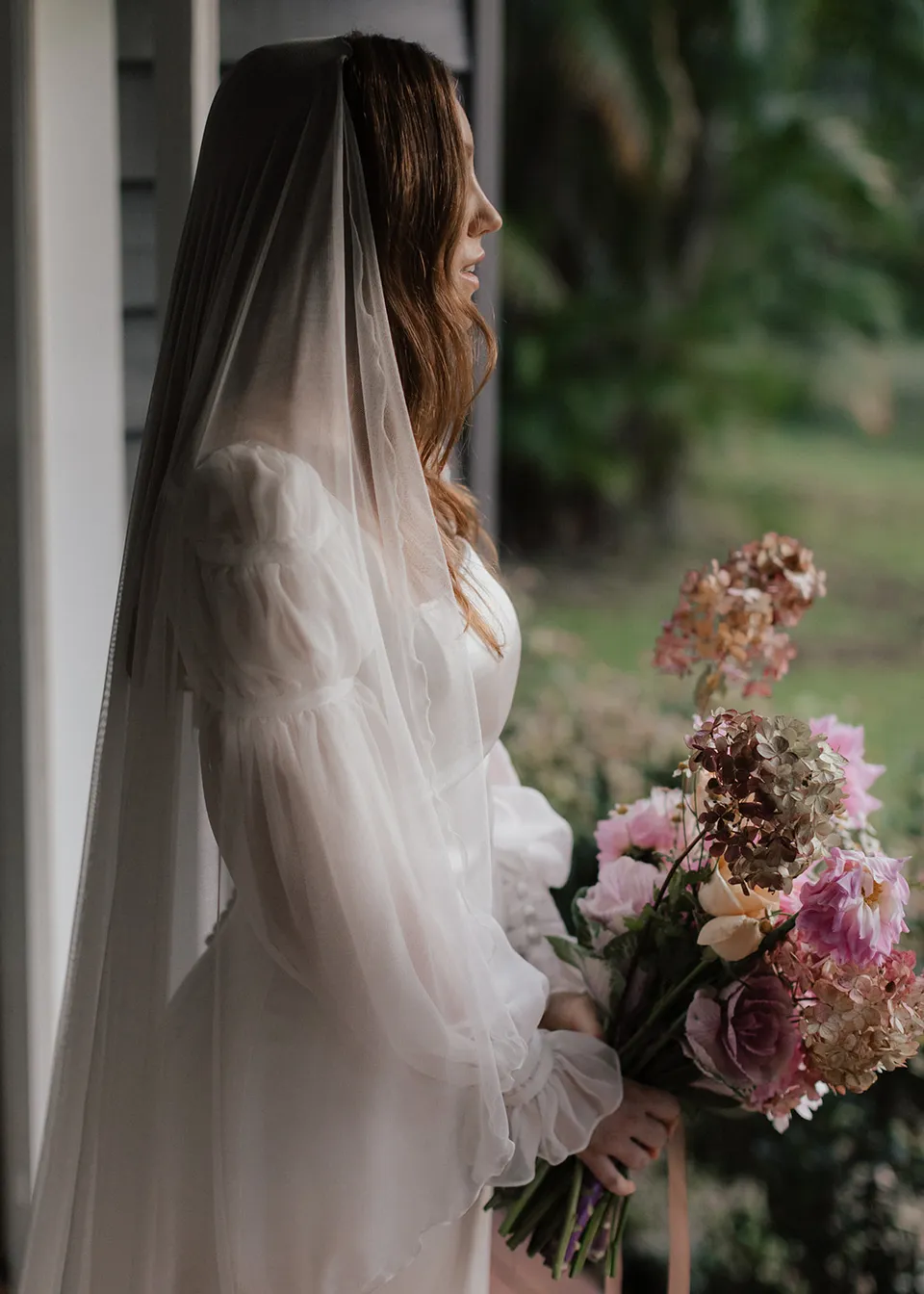 Side portrait of Elyse in her veil holding a pink bouquet by the doorway, with flowers by The Perfect Party Co. At Fig Tree Villa in Ewingsdale, Byron Bay, for an Elopement Collective elopement. Photograph by Bulb Creative.
