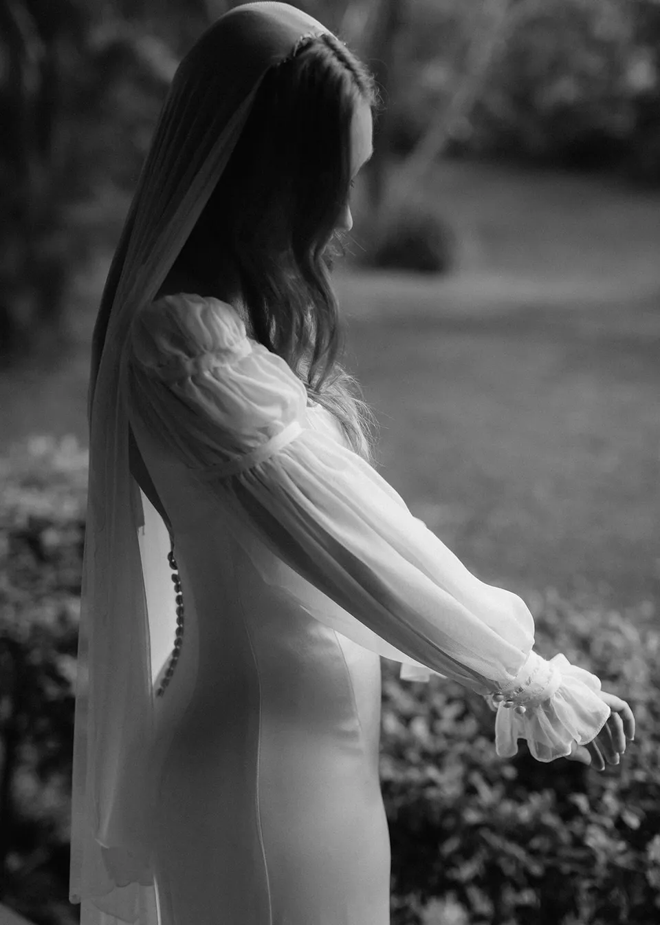 Black-and-white photograph of Elyse looking down at her sleeves outdoors. At Fig Tree Villa in Ewingsdale, Byron Bay, for an Elopement Collective elopement. Photograph by Bulb Creative.