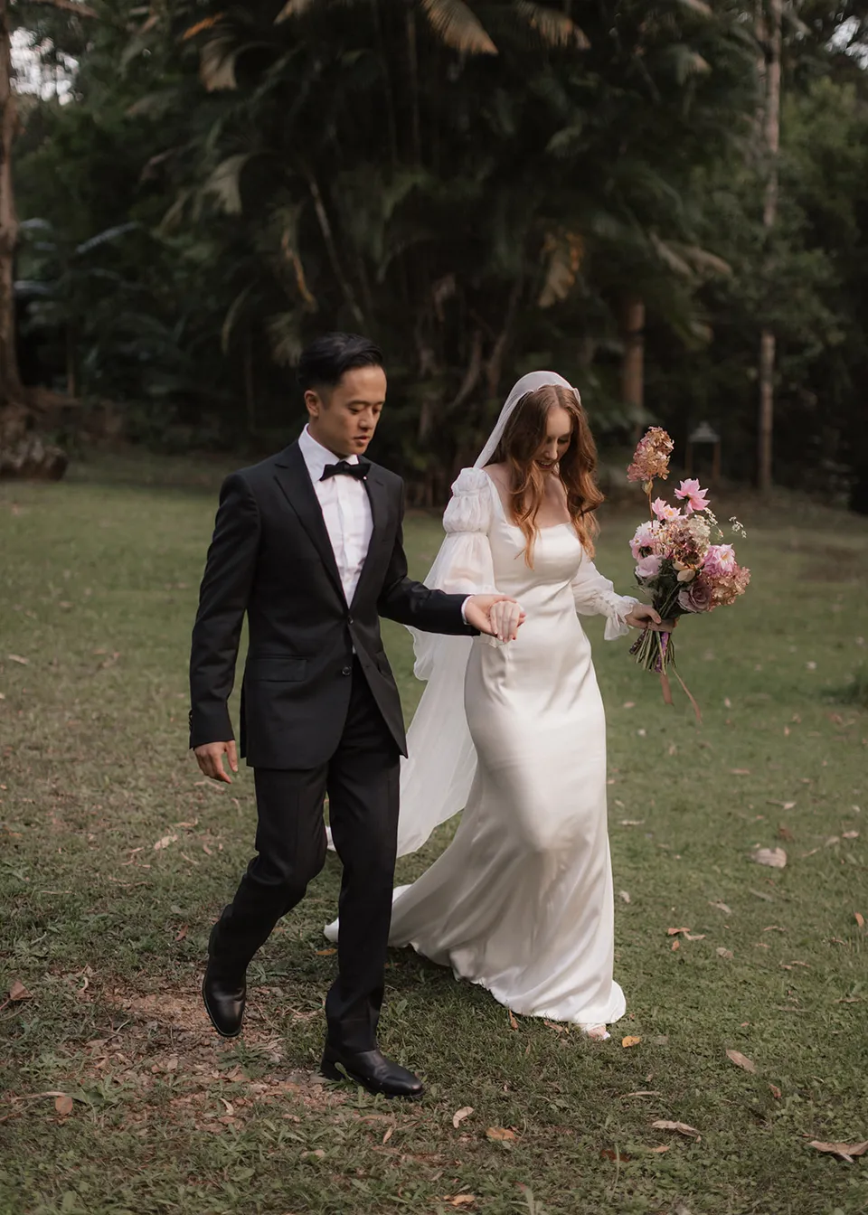 Elyse and Jonathan walking hand in hand across the lawn with her bouquet, with flowers by The Perfect Party Co. At Fig Tree Villa in Ewingsdale, Byron Bay, for an Elopement Collective elopement. Photograph by Bulb Creative.