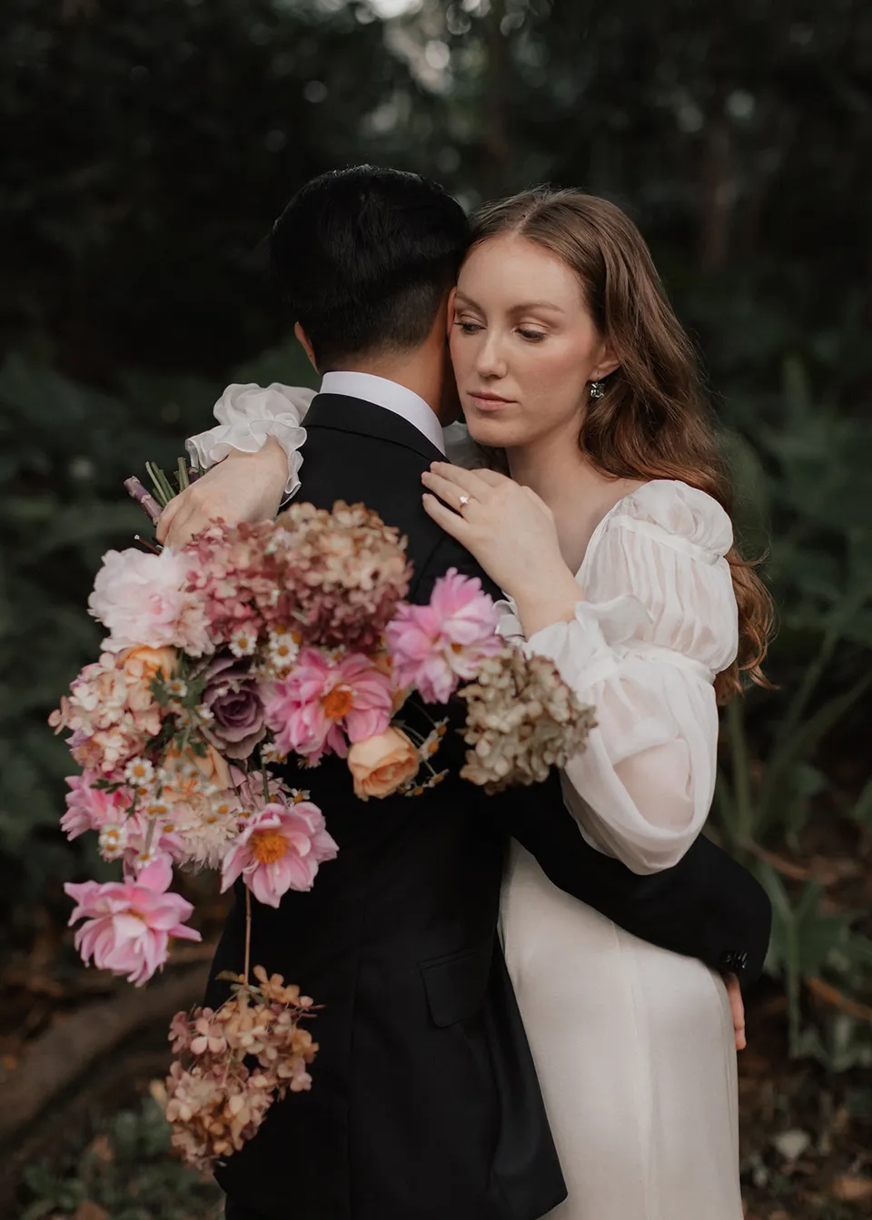 Elyse resting against Jonathan with her pink bouquet over his shoulder, with flowers by The Perfect Party Co. At Fig Tree Villa in Ewingsdale, Byron Bay, for an Elopement Collective elopement. Photograph by Bulb Creative.