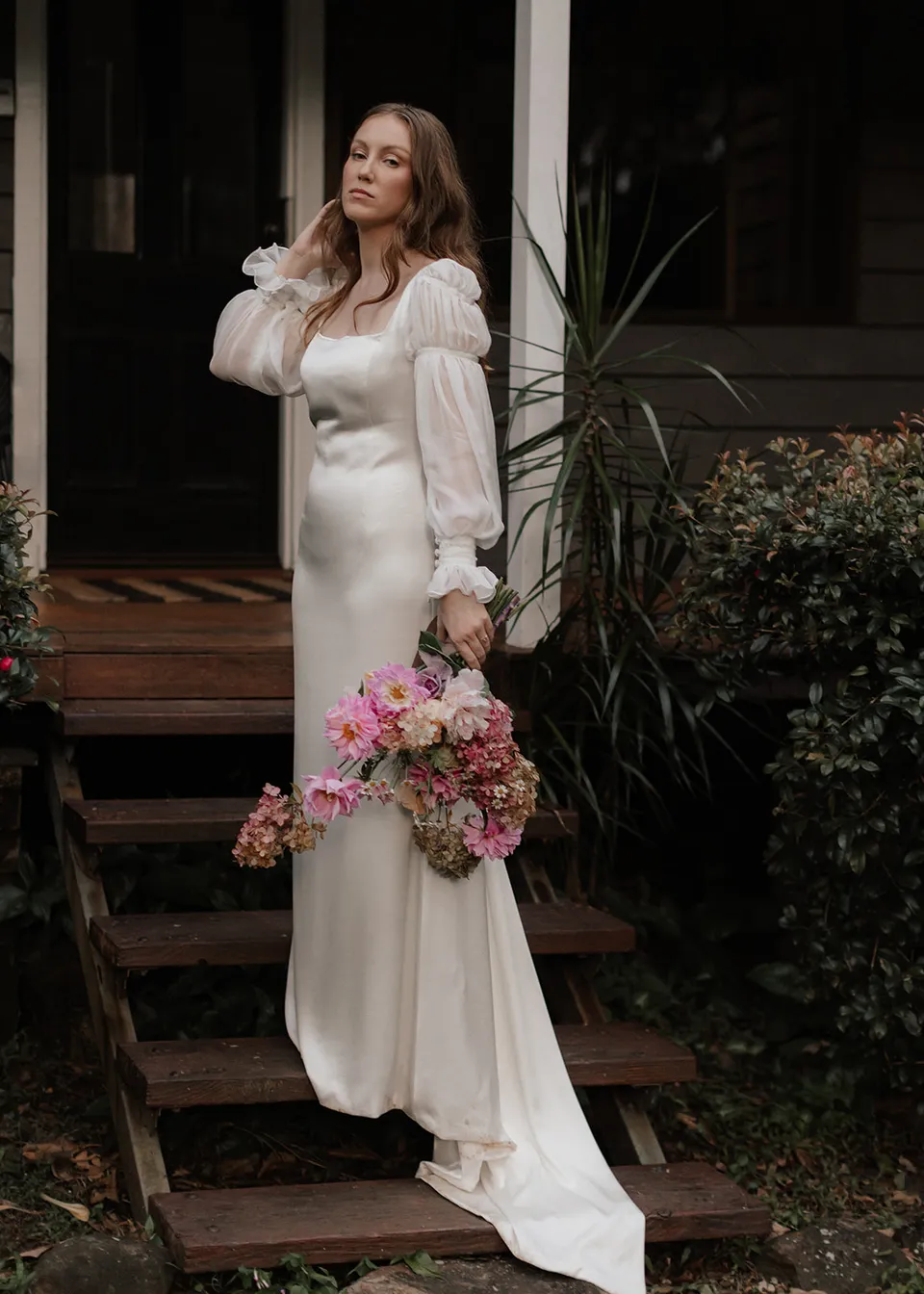Elyse brushing back her hair beside the cottage steps. At Fig Tree Villa in Ewingsdale, Byron Bay, for an Elopement Collective elopement. Photograph by Bulb Creative.