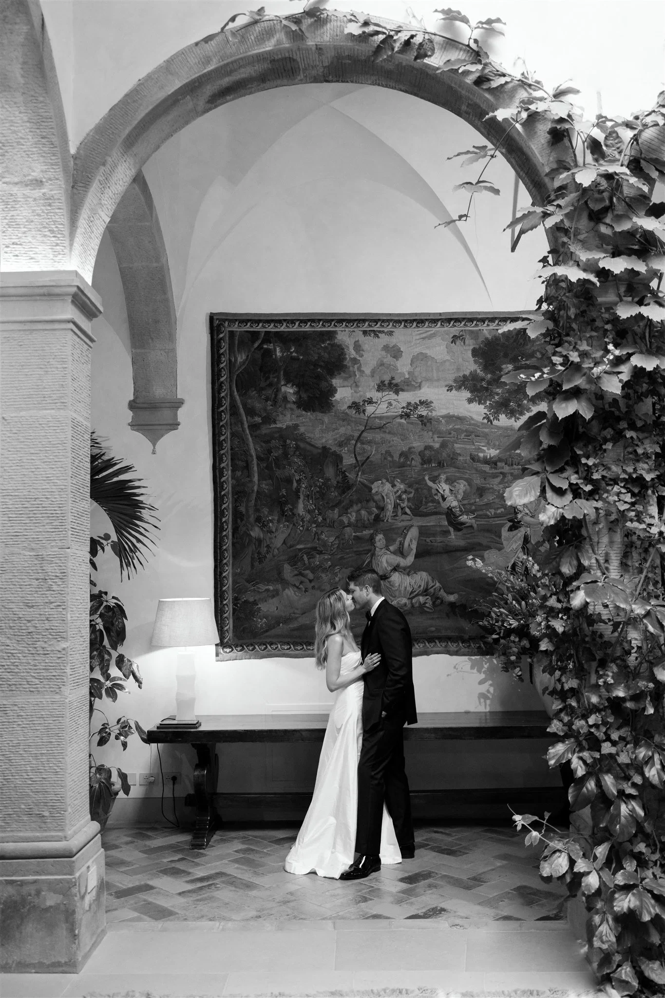 Black-and-white photograph of Holly and Matt kissing in a quiet arcaded courtyard framed by leafy vines, at Belmond Villa San Michele in Fiesole, overlooking Florence, Tuscany, Italy, for an Elopement Collective elopement. Photo by Khara from House of Lucie.