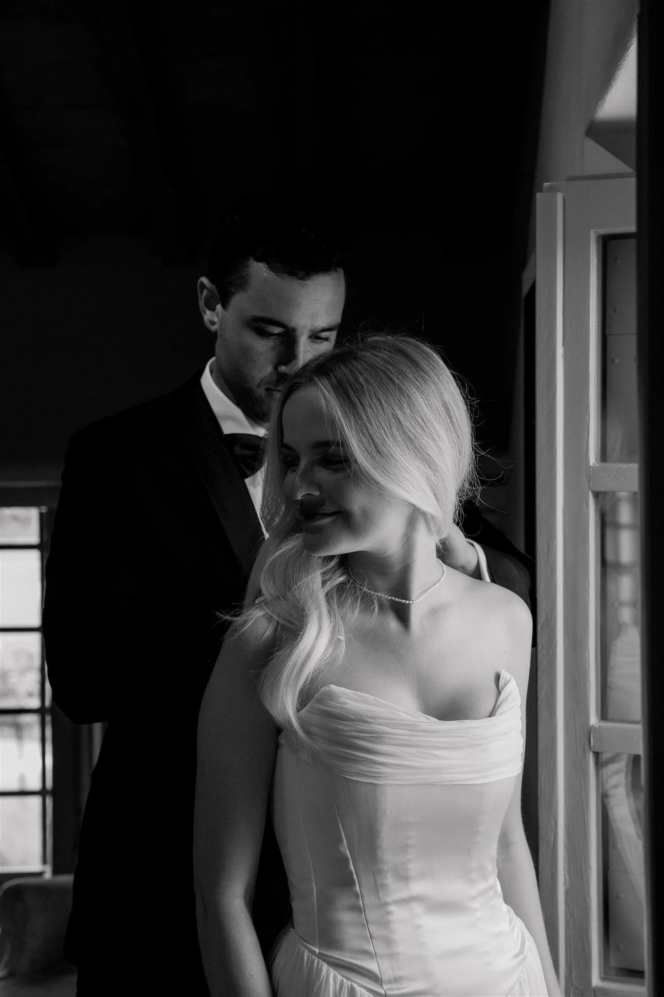Black-and-white photograph of Jack kissing Shelby’s head as she stands beside an open window, in Umbria, Italy, for an Elopement Collective elopement. Photo by James from House of Love.