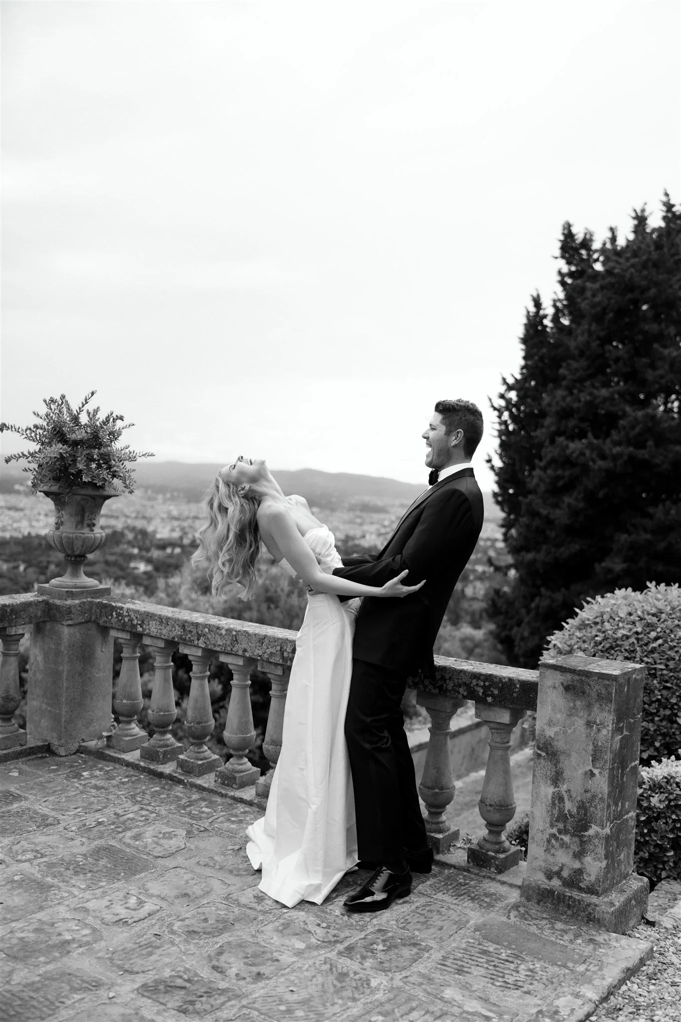 Black-and-white photograph of Matt leaning back as he holds Holly close beside the balustrade, at Belmond Villa San Michele in Fiesole, overlooking Florence, Tuscany, Italy, for an Elopement Collective elopement. Photo by Khara from House of Lucie.