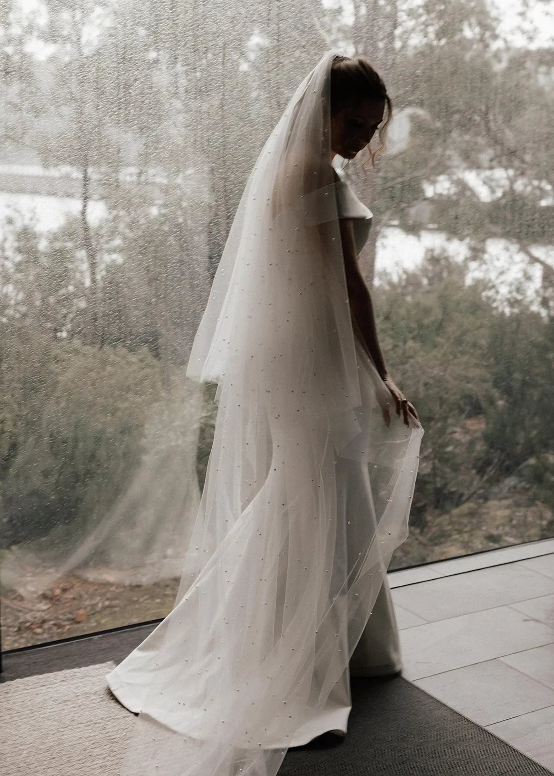 Sarah looking down at her white dress and long pearl-dotted veil beside the window, at Pumphouse Point, Lake St Clair, Tasmania, for an Elopement Collective elopement. Photo by Bulb Creative.
