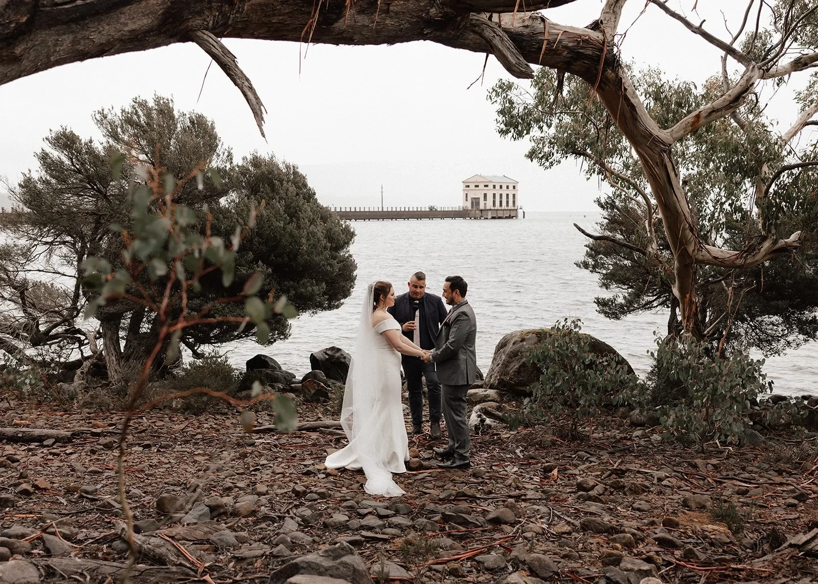 Sarah and Jono holding hands during their lakeside ceremony with celebrant Josh Withers beneath spreading trees, at Pumphouse Point, Lake St Clair, Tasmania, for an Elopement Collective elopement. Photo by Bulb Creative.