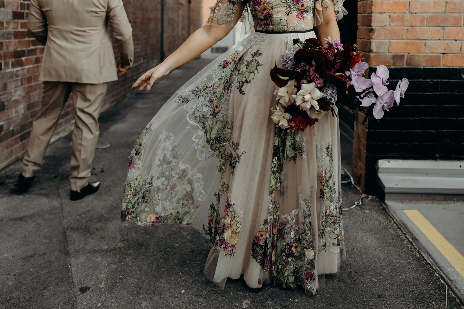 Victoria lifting the sheer floral layer of her skirt while holding her bouquet by Mrs Gibbons Flowers, with Daniel walking ahead, in Fortitude Valley, Brisbane, for an Elopement Collective elopement. Photo by Tessa Shannon.