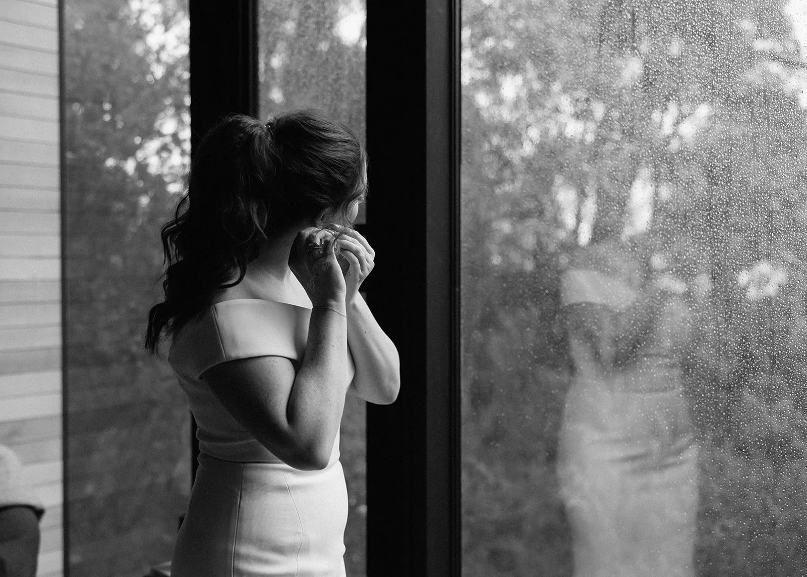 Black-and-white photograph of Sarah adjusting an earring beside a large window, at Pumphouse Point, Lake St Clair, Tasmania, for an Elopement Collective elopement. Photo by Bulb Creative.