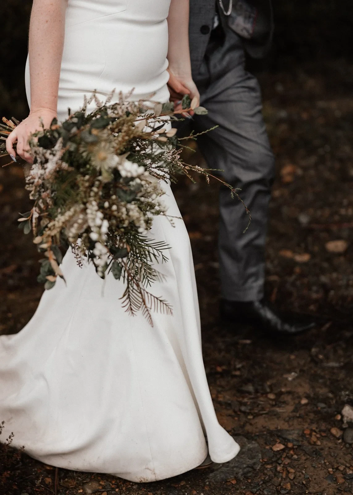 Close-up of Sarah’s bouquet by The Romantics against her white skirt beside Jono, at Pumphouse Point, Lake St Clair, Tasmania, for an Elopement Collective elopement. Photo by Bulb Creative.