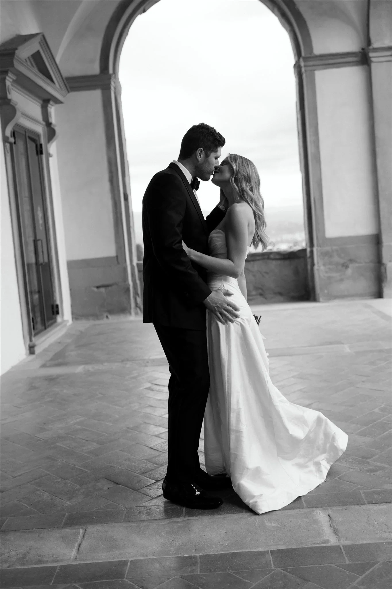 Black-and-white photograph of Matt dipping Holly into a kiss beneath a stone arcade, at Belmond Villa San Michele in Fiesole, overlooking Florence, Tuscany, Italy, for an Elopement Collective elopement. Photo by Khara from House of Lucie.