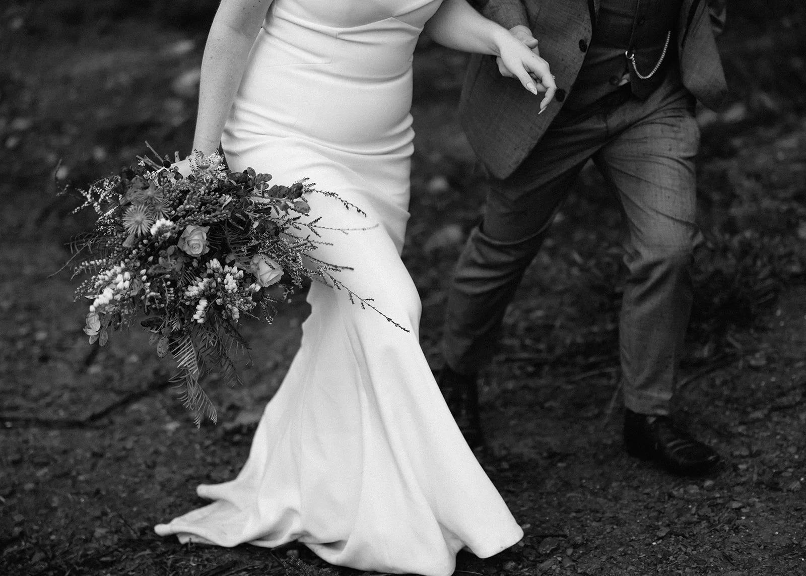 Black-and-white detail of Sarah gathering her dress and holding her bouquet by The Romantics while linked to Jono’s arm, at Pumphouse Point, Lake St Clair, Tasmania, for an Elopement Collective elopement. Photo by Bulb Creative.